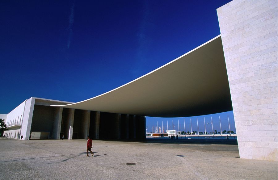 Amplio pabellón con techo ondulado blanco, sostenido por pilares, sobre una explanada. Una persona de rojo camina por el espacio abierto. Fondo con mástiles y vegetación.