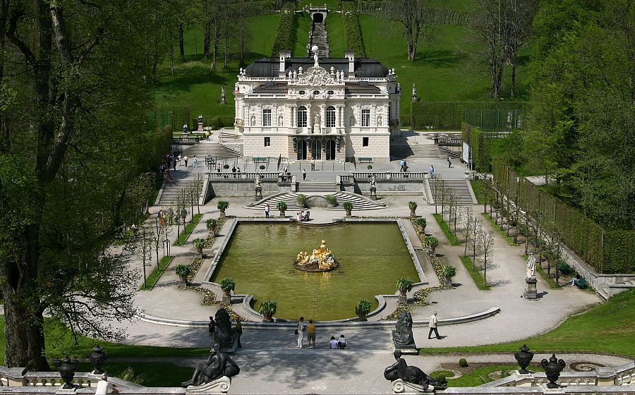 Vista panorámica del Palacio de Linderhof: arquitectura blanca, jardines geométricos, fuente dorada, visitantes y cascada artificial.
