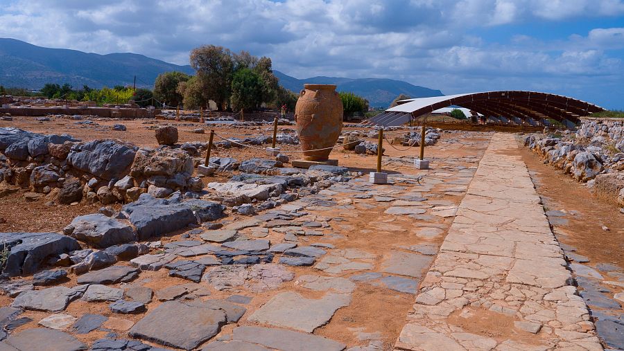Restos arqueológicos con camino empedrado, vasija de cerámica en primer plano y estructura moderna de protección al fondo. Paisaje montañoso al horizonte.