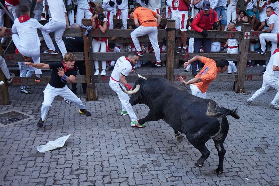 Toro negro corriendo en calle estrecha durante un encierro. Dos corredores lo esquivan; uno viste camiseta blanca y otro naranja.  Espectadores observan desde vallas altas.