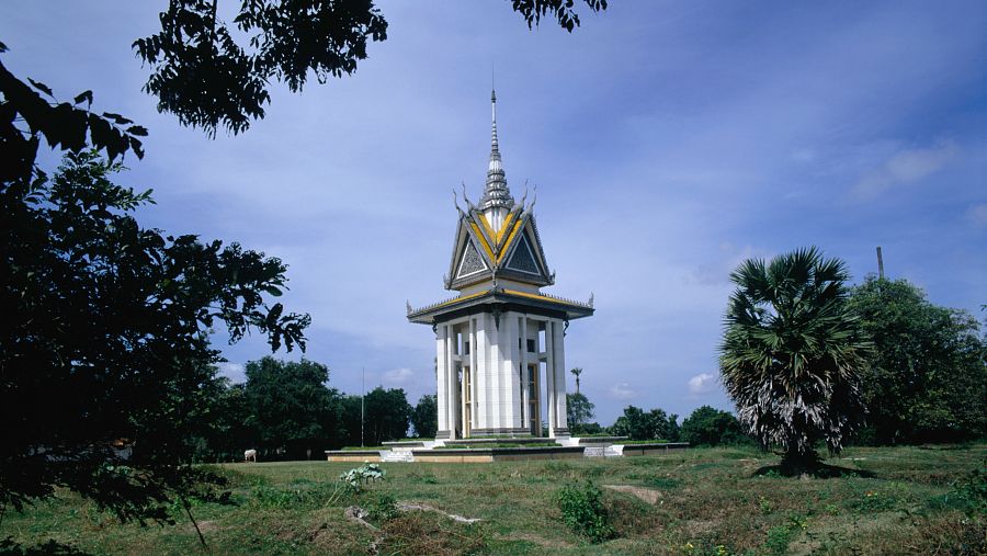 Monumento jemer de color blanco y dorado en un espacio abierto con vegetación.  Presenta una estructura alta con varios niveles y montículos de tierra en primer plano.