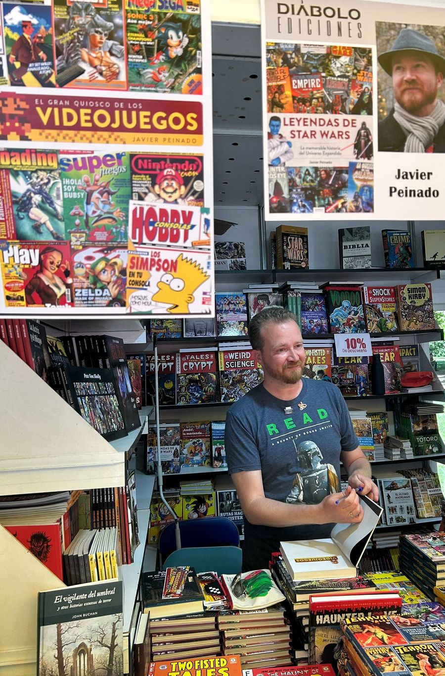 Stand en una feria del libro con Javier Peinado firmando libros.  Se exhiben cómics, libros y material de videojuegos retro, con carteles promocionales de títulos clásicos.