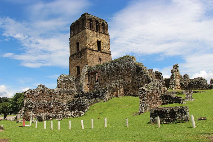 Ruinas de piedra de una catedral, con una torre principal parcialmente conservada y vegetación entre los restos.  Cielo azul y postes blancos en primer plano.