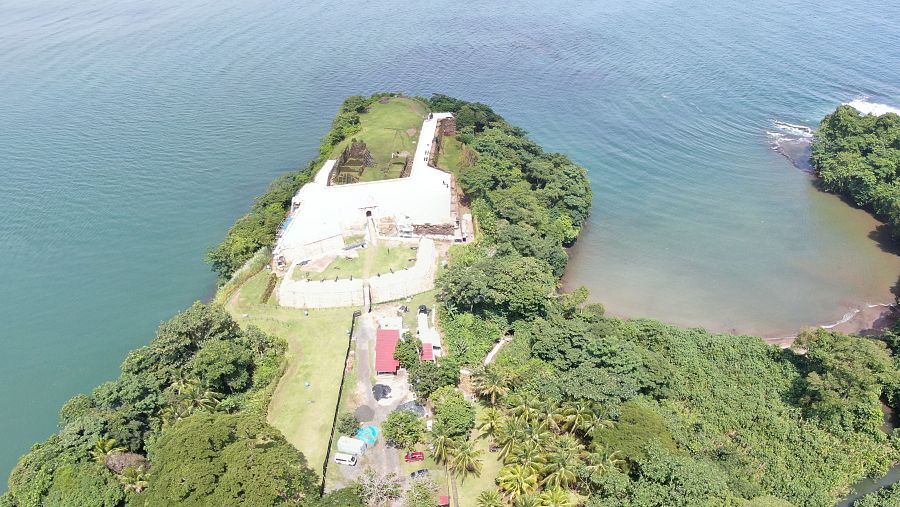 Impresionante vista aérea de una fortaleza blanca en un promontorio, rodeada de agua y vegetación. Edificios menores visibles cerca.