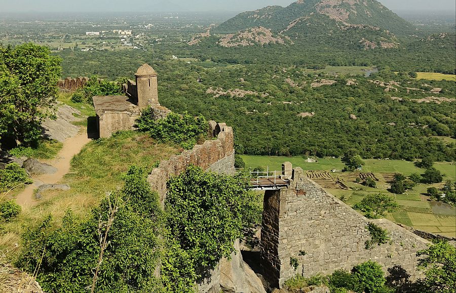 Vista de las murallas de una fortificación histórica, con un torreón, puente metálico y vegetación. El paisaje incluye colinas, bosques y campos.
