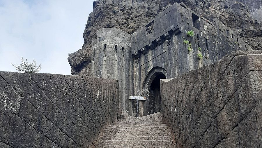 Empinada escalera de piedra conduce a un arco de piedra oscuro, entrada a un fuerte.  Muros de piedra inclinados flanquean el estrecho pasillo.  El camino muestra desgaste.