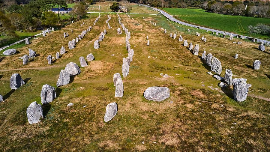 Vista aérea de un alineamiento megalítico con menhires de diversos tamaños en filas sobre terreno irregular con vegetación seca y una carretera al fondo.