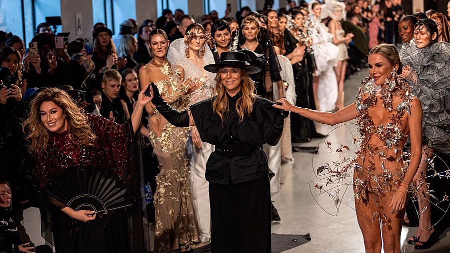 Diseñadora con sombrero negro y atuendo oscuro saluda tras un desfile de alta costura, acompañada de una cantante con vestido burdeos.