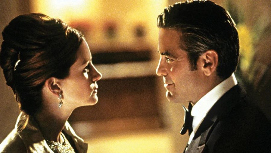 Intensa mirada entre Julia Roberts, con moño alto y vestido claro, y George Clooney, con esmoquin negro.  Posible escena de película, fondo borroso.