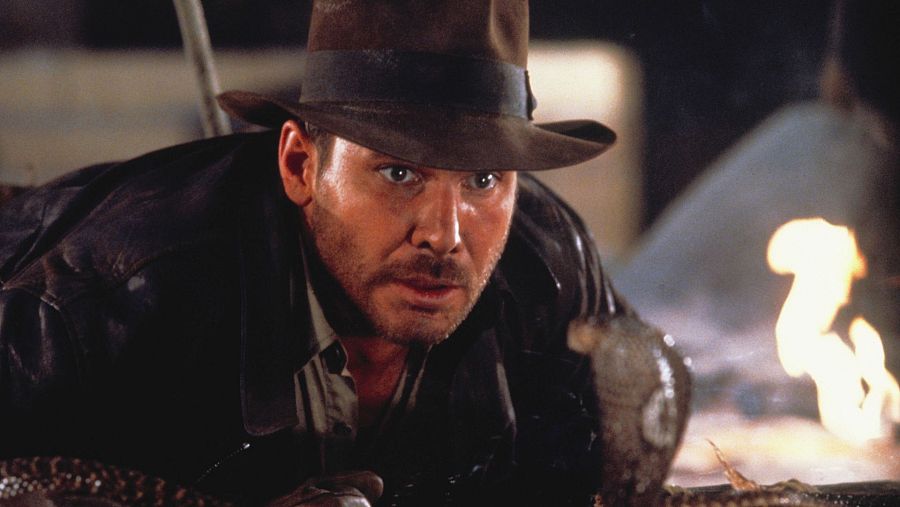 Indiana Jones, agachado en la oscuridad, con una serpiente cerca de su cara y una llama al fondo.  Su expresión denota miedo y cautela.