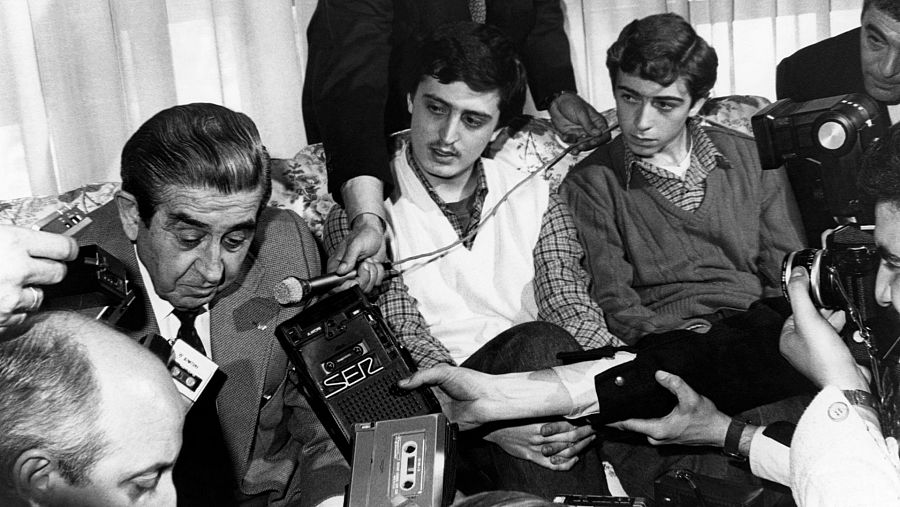 Rueda de prensa tras liberación. Empresario en camisa a cuadros y chaleco responde a periodistas con micrófonos y grabadoras.  Ambiente doméstico y tenso.