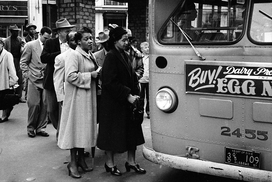 Rosa Parks y otras personas esperan subir a un autobús en una calle. Imagen en blanco y negro de la década de 1950, con un anuncio y parte de la matrícula del autobús visible.