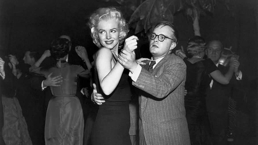 Marilyn Monroe y Truman Capote bailando en un club nocturno; ella con vestido oscuro y él con traje a rayas y gafas.  Ambiente festivo.
