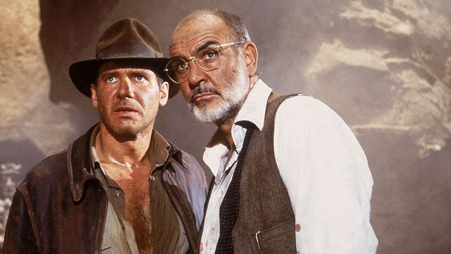 Indiana Jones y su padre, Henry Jones Sr., con expresiones serias, observan algo fuera de cuadro en un ambiente oscuro.
