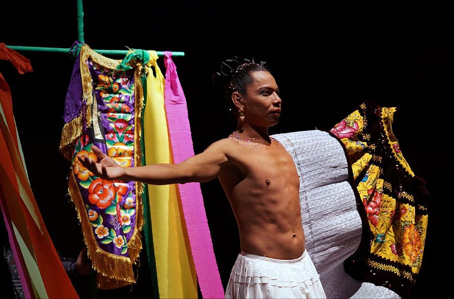 Bailarín con falda blanca y torso desnudo, en pose expresiva, frente a un fondo de telas coloridas con bordados y flecos.