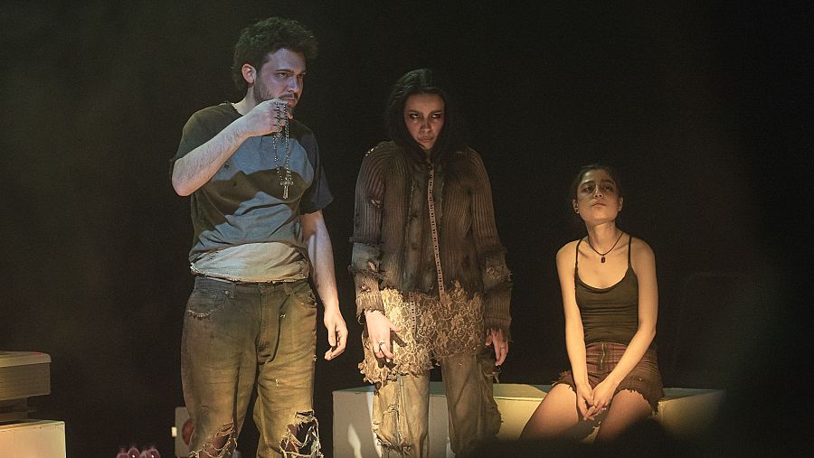 Tres actores en escena con ropa desgastada, interpretando una obra dramática con fondo oscuro y dos cajas blancas.  Expresiones serias.