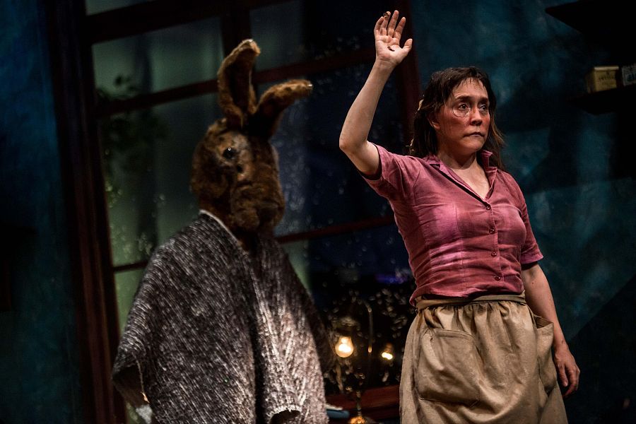 Escena teatral: personaje enmascarado de conejo y mujer con blusa rosa y falda beige interactúan en un escenario oscuro con iluminación baja.  Ambiente dramático.
