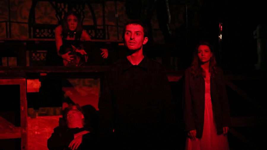 Intensa escena teatral con iluminación roja, mostrando a un hombre mirando hacia arriba, una mujer de pie y otra en una plataforma elevada; un actor yace en el suelo.