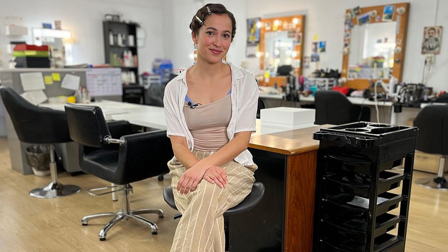 Mujer con el pelo recogido, camiseta beige, camisa blanca y pantalón beige a rayas, sentada en un taburete en una sala de maquillaje con micrófono inalámbrico.