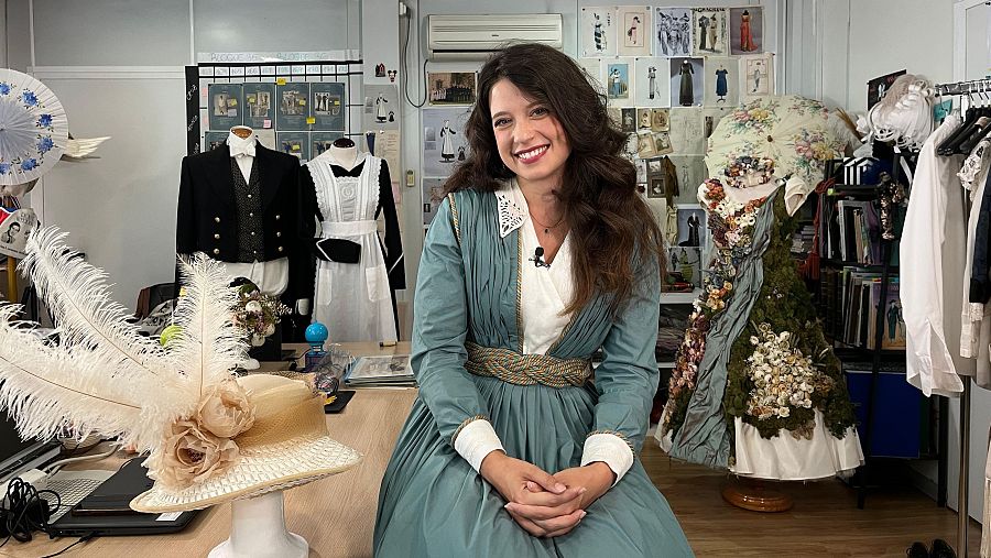 Actriz con vestido verde azulado en vestuario de rodaje.  Se aprecian trajes, bocetos y accesorios. Ambiente de trabajo creativo.