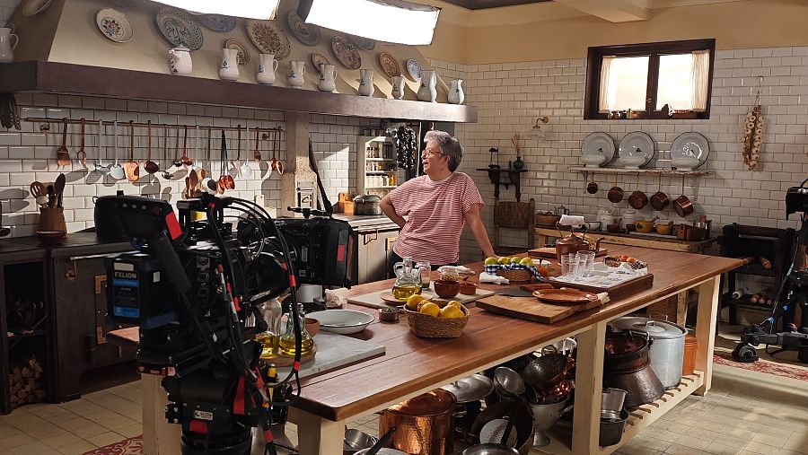 Eva Bermúdez de Castro en la cocina de un rodaje, con una mesa de madera, utensilios de cocina antiguos y equipo de grabación visible.