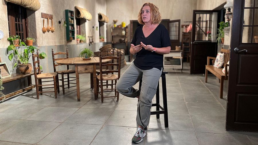 Operadora de cámara en el set de rodaje, sentada en un taburete, con ropa informal y gesticulando.  Ambiente rústico con muebles de madera y plantas.