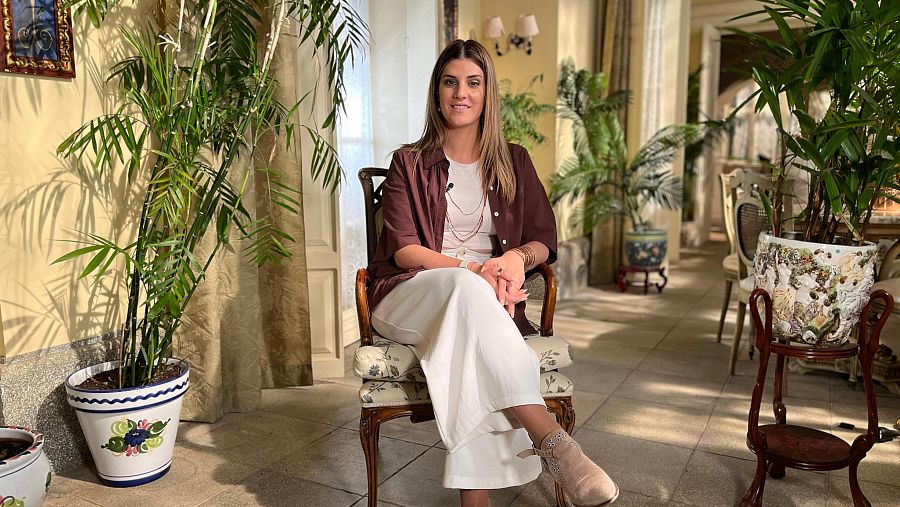 Mujer con camisa marrón, pantalones blancos y botas beige sentada en silla de madera en galería iluminada con plantas. Lleva micrófono y collares.