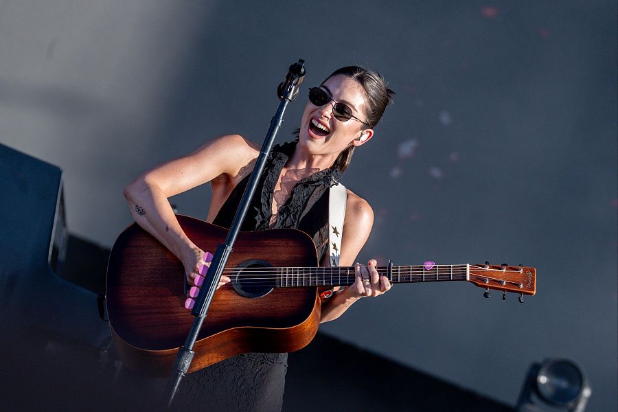 Cantante con gafas de sol tocando guitarra acústica en un escenario con fondo gris, sonriendo y con púas rosas en los dedos.