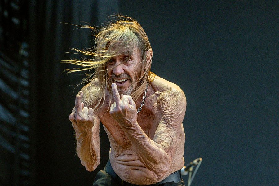 Iggy Pop, torso desnudo y cabello rubio despeinado, muestra enfado con gestos y expresión facial durante un concierto, aparentemente por fallos técnicos.