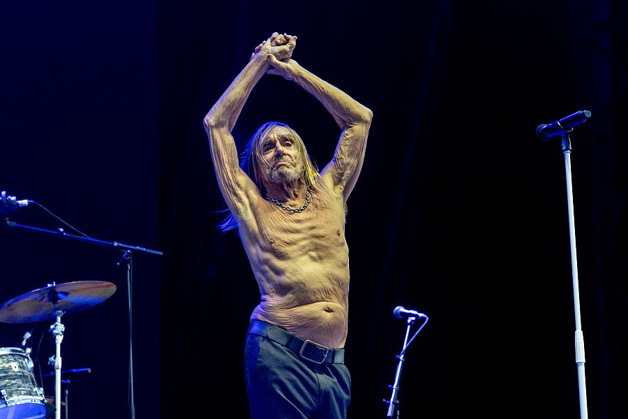 Imagen de Iggy Pop en concierto: sin camisa, con cadena y brazos en alto, expresión seria. Fondo oscuro con elementos del escenario.
