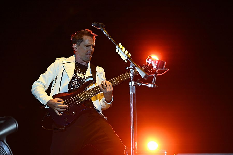Matthew Bellamy, con chaqueta blanca y camiseta oscura, toca la guitarra en un escenario iluminado con luces anaranjadas. Se ve un micrófono.
