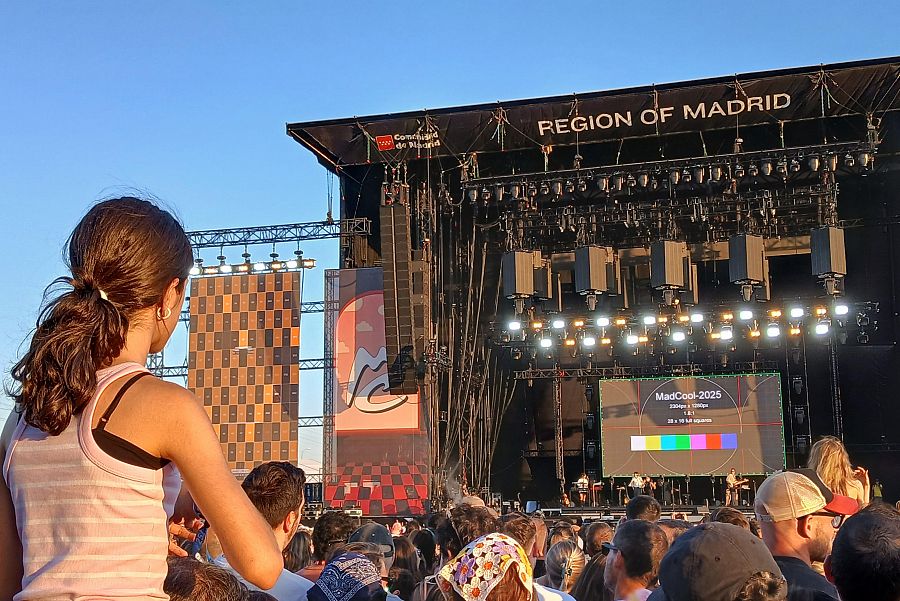 Niña en hombros observa pantalla gigante con imagen de prueba en festival musical, concierto interrumpido por problemas técnicos.