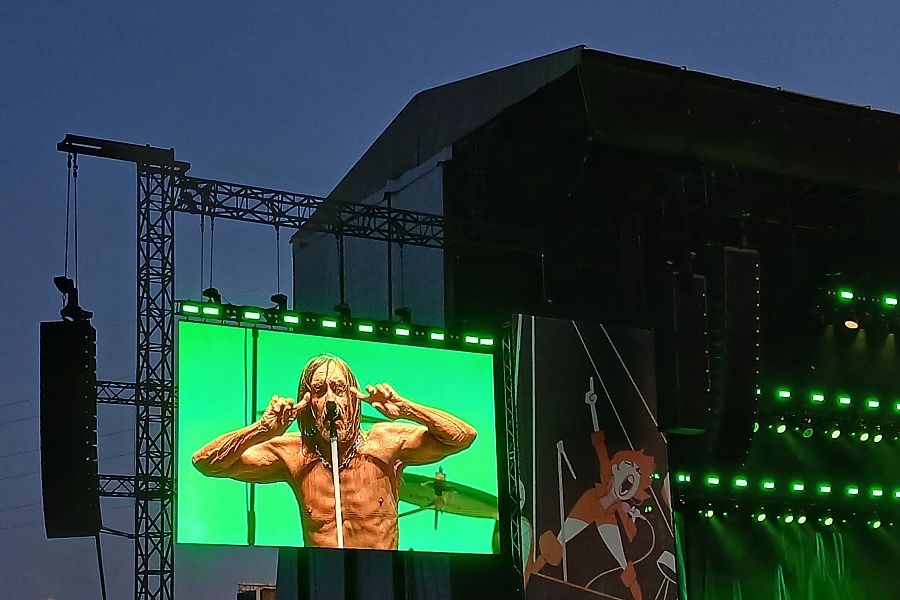 Imagen de Iggy Pop en pantalla gigante durante un concierto.  Sin camiseta, con el pelo rubio y haciendo un gesto con las manos. Fondo verde brillante en contraste con el escenario.