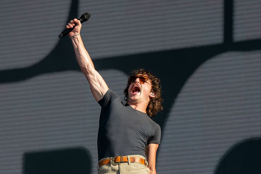 Cantante con gafas de sol y micrófono, brazo derecho alzado, cantando enérgicamente sobre un fondo oscuro. Concierto en Madrid.
