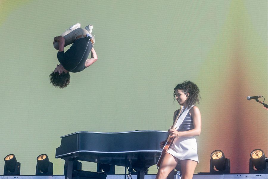 Cantante realizando un salto mortal en un concierto, con una guitarrista y un piano de cola en el escenario.