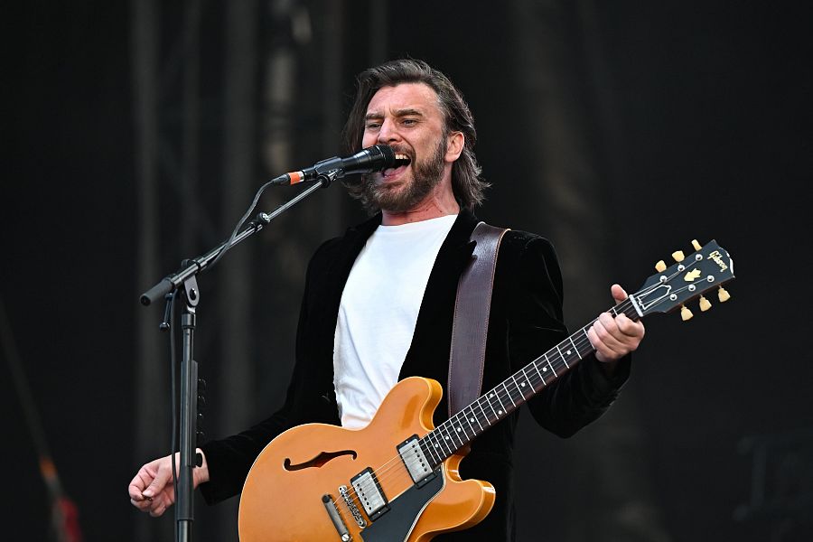 Vocalista de rock en concierto, cantando al micrófono y tocando una guitarra Gibson ES-335.  Chaqueta oscura, pelo castaño y expresión concentrada.