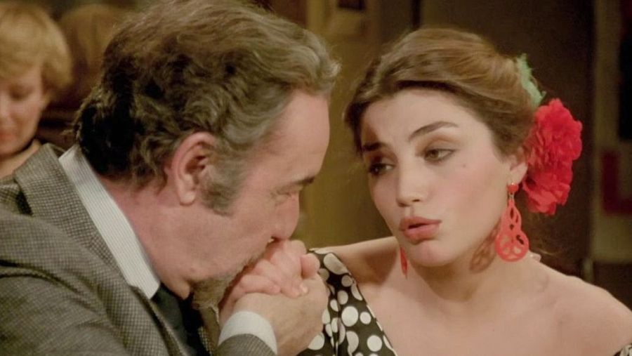 Escena de interior: hombre mayor besa la mano de una mujer con vestido de lunares y flor roja en el pelo.  Otra persona al fondo.