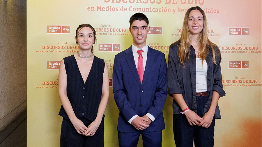 Tres personas en traje formal posan ante un cartel amarillo que anuncia una jornada sobre discursos de odio en medios y redes sociales, posiblemente relacionada con RTVE.