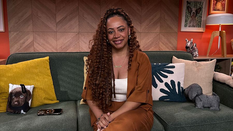 Mujer sentada en sofá verde con cojines, teléfono móvil cercano.  Ambiente acogedor con temática canina, decorado con cuadros de perros y lámpara de pie.