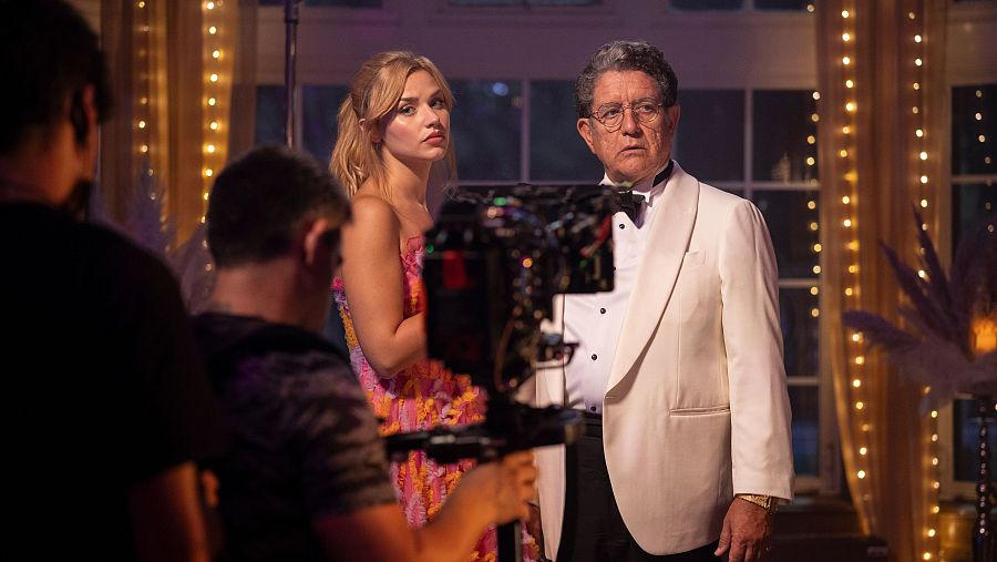 Rodaje de película: actores (mujer con vestido rosa y naranja, hombre con esmoquin blanco) interpretan una escena en un decorado elegante, grabada con cámara profesional.