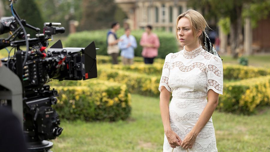 Actriz con vestido blanco y lazo negro en el pelo, en rodaje con cámara y equipo de filmación en jardín.  Ambiente de época o clase alta.