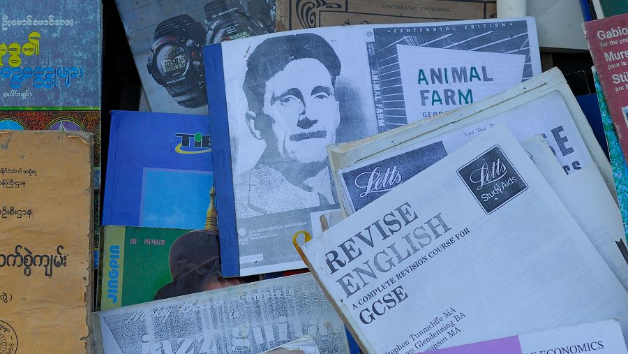 Pila de libros y folletos en un puesto, con títulos en inglés y birmano, incluyendo 