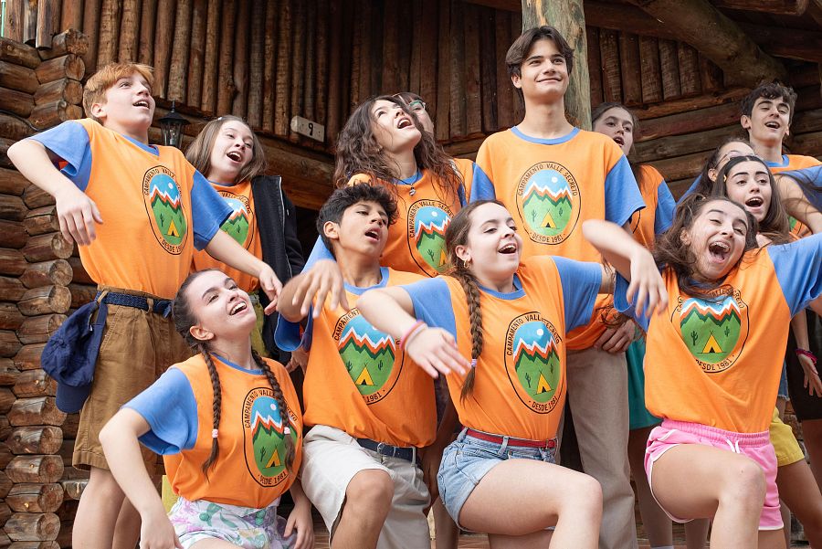 Niños y adolescentes sonríen y posan con entusiasmo frente a una cabaña, vistiendo camisetas con un logo de campamento. Actividad grupal en un entorno campestre.