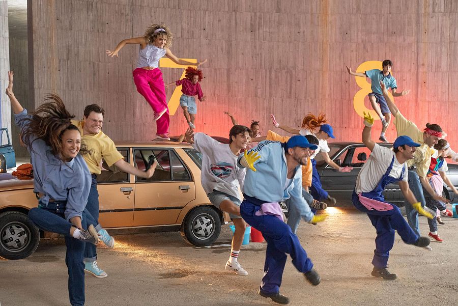 Bailarines con ropa de los 80 en una escena vibrante sobre coches en un aparcamiento o túnel de lavado.  Colores brillantes y mucha energía.