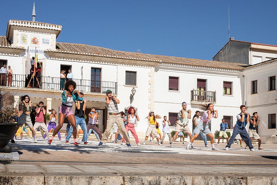 Jóvenes con ropa de estilo ochentero bailan en una plaza frente a un edificio de dos plantas con balcones, donde se ven personas observando.
