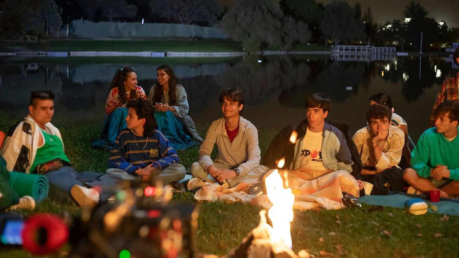 Grupo de jóvenes alrededor de una fogata en la noche, junto a un lago.  Ambiente oscuro, iluminado por el fuego. Se ve equipo de filmación.