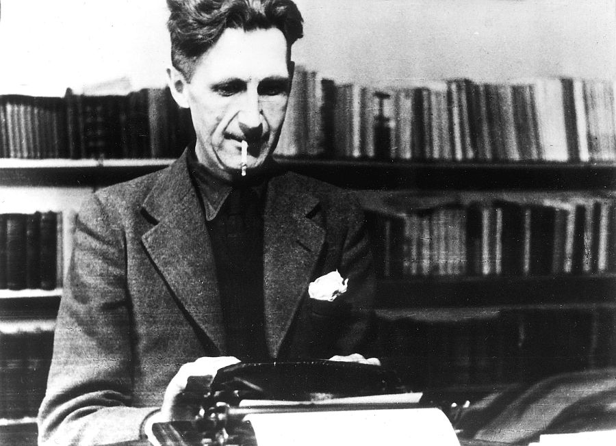 Orwell, concentrado en su máquina de escribir, fuma un cigarrillo.  Viste americana y camisa oscura;  al fondo, una estantería repleta de libros.