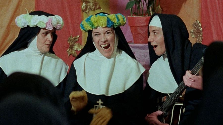 Tres monjas en escena musical dentro de un convento.  Una canta, otra lleva una corona de flores y la tercera toca la guitarra.  Escena festiva con contraste entre atuendo religioso y música.