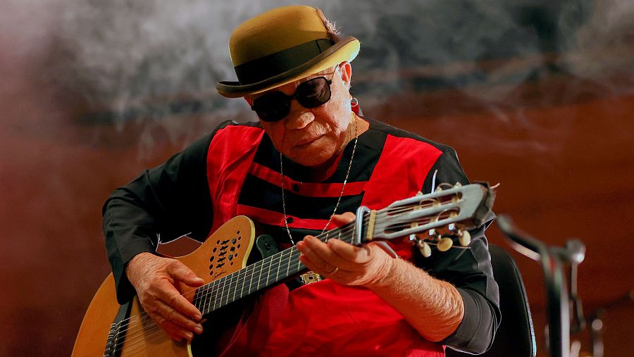 Músico tocando la guitarra en un escenario con humo; viste camisa roja, sombrero beige y gafas de sol.