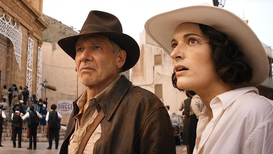 Harrison Ford y Phoebe Waller-Bridge, con sombreros, miran hacia arriba en una escena de 'Indiana Jones y el dial del destino'.  Se observa una plaza con un edificio y personas al fondo.